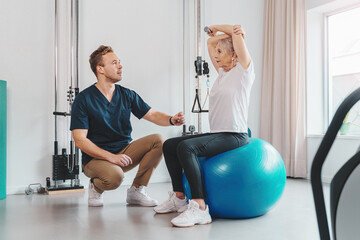 Caucasian senior woman sitting on a Pilates ball, her personal trainer, kneeling down, gives her...