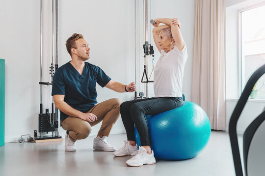 Caucasian senior woman sitting on a Pilates ball, her personal trainer, kneeling down, gives her instructions. They're in a gym, smiling, exercising together.