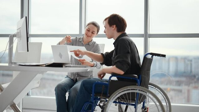 Young male office worker in a wheelchair talking to female colleague while showing her ideas using laptop in co-working space. Disability and handicap concept. Office modern people at work.
