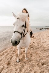 A white horse and a woman in a dress stand on a beach, with the sky and sea creating a picturesque backdrop for the scene.