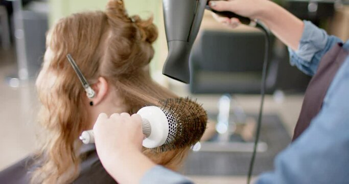 Caucasian female hairdresser styling client's long hair with hairdryer and brush, in slow motion