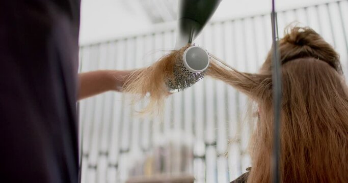 Caucasian female hairdresser styling client's long hair with hairdryer and brush, in slow motion
