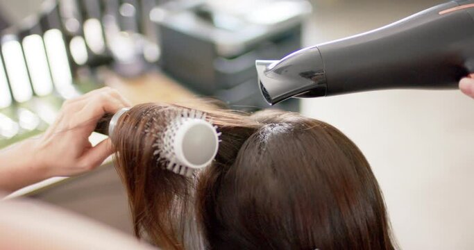 Caucasian female hairdresser styling client's long hair with hairdryer and brush, slow motion