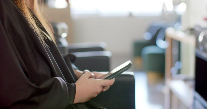Midsection of caucasian female client in cape at hair salon using smartphone, in slow motion