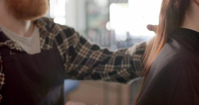 Bearded caucasian male hairdresser combing client's long hair at salon, in slow motion