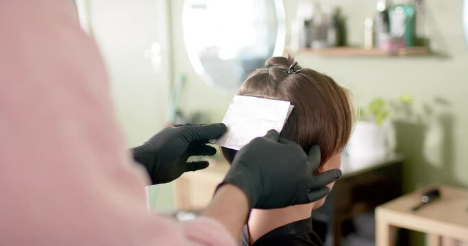 Hands of caucasian male hairdresser highlighting client's hair in foil at salon, in slow motion