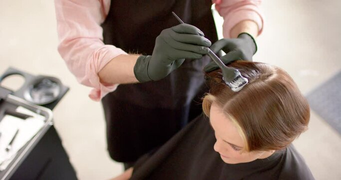Midsection of caucasian male hairdresser dyeing happy female client's hair at salon, in slow motion