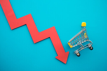 Shopping trolley with red chart falling down on blue background copy space. Economic recession crisis, core retail sales decrease, inflation or goods price up concept.