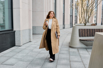 The urban landscape, businesswoman adorned in a fashionable trench coat, navigates the city's streets with her laptop and a cup of aromatic coffee in hand