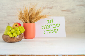 Bouquet of wheat ears, notebook paper with Hebrew text, fresh fruits for Jewish holiday Shavuot on white wooden background. Hebrew text on notebook paper