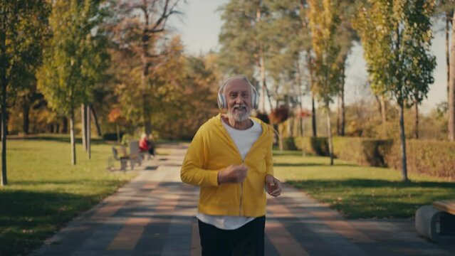 Happy senior man in headset jogging in sunny park, enjoying morning run, sport