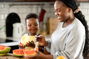 Cheerful African-American mother and son eating a tasty watermelon at home. Flavors of summer