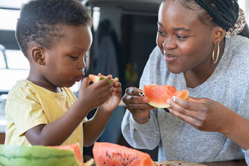 Cheerful African-American mother and son eating a tasty watermelon at home. Flavors of summer