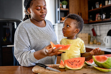 Cheerful African-American mother and son eating a tasty watermelon at home. Flavors of summer