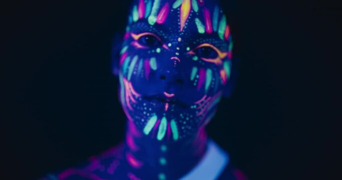 Beauty Close Up Footage: Young Female Poses with Confidence, Neon Paint Accentuating Her Facial Features Against an Low Key Studio Setting, Creating an Artistic Appearance