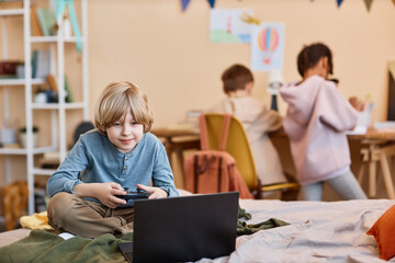 Portrait of blonde preteen boy playing videogame on bed with laptop, copy space