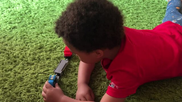 Three-year-old black toddler wearing a red shirt playing with a toy train lying on a green rug.