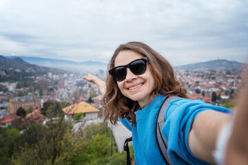 Young happy woman in sunglasses taking selfie with view of Bosnian capital Sarajevo, travel to Balkans