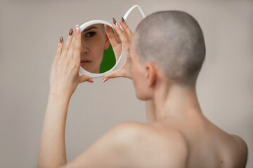 a young slender woman with a shaved head stands with her bare back to us and examines herself in the mirror, selective focus. Side effects of chemotherapy in cancer treatment