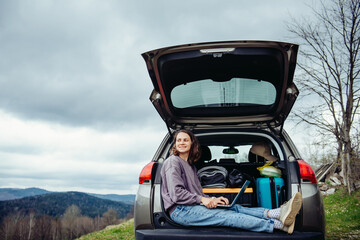 Young pretty smiling woman traveler in casual wear sitting in car trunk using laptop while...