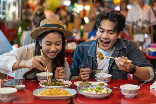 Young Asian couple traveler tourists eating Thai street food together in China town night market in Bangkok in Thailand - people traveling enjoying food culture concept