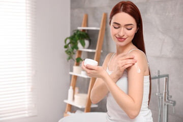 Beautiful young woman applying body cream onto shoulder in bathroom, space for text