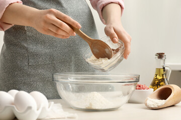 Woman putting flour into bowl at white wooden table, closeup