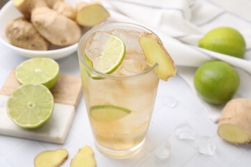 Glass of tasty ginger ale with ice cubes and ingredients on white tiled table, closeup