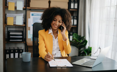 Obraz premium Business African woman Talking on the phone and using a laptop with a smile while sitting at office