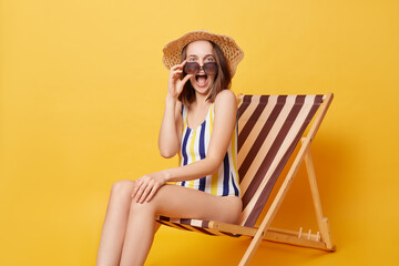 Overjoyed delighted young woman wearing striped swimwear and hat sitting on deck chair isolated over yellow background screaming with excitement looking at camera rejoicing her vacation.