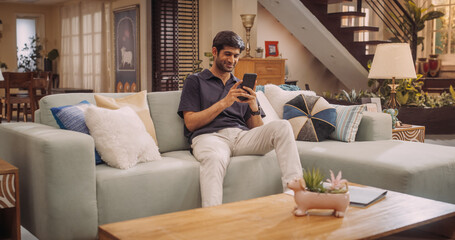 Cheerful Young Hindu Man Comfortably Using His Smartphone At Home, Handsome, Smiling and Positive. He's Scrolling Through Social Media Or Engaging In Virtual Remote Work. 