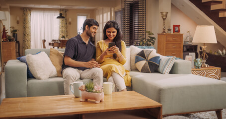 Young Indian Couple Using Internet On Smartphone, Sitting On The Sofa at Home, they Tease and Joke...