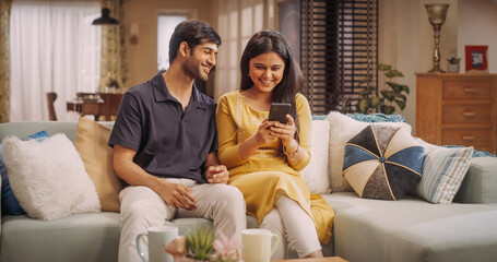 Young Indian Couple Using Internet On Smartphone, Sitting On The Sofa at Home, they Tease and Joke...