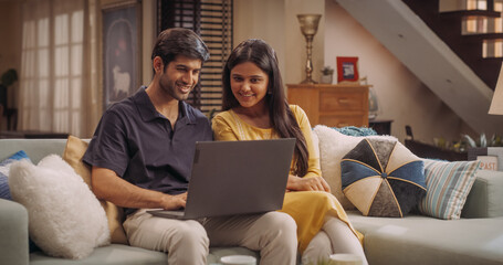 Adorable Young Indian Couple Using Laptop, Sitting on Home Sofa. Exploring Online Content, Discussing Trend Ideas and Making Purchase Decisions Together. Online Shopping Concept. 
