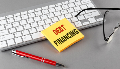 DEBT FINANCING text on a sticky with keyboard, pen glasses on grey background