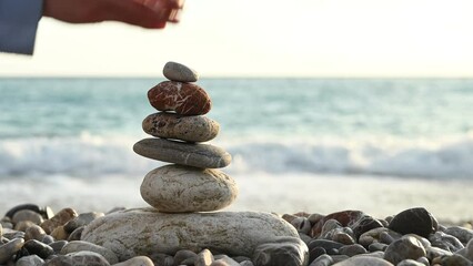 Unrecognizable woman trying to build balanced rock pyramid, but fails. Breaking balanced stones on pebbles beach against sea waves on background - Powered by Adobe