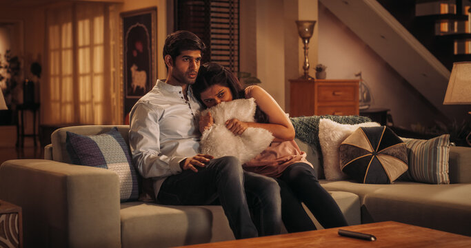 
Young Indian Couple Watching Scary Movie on TV, Finds Comfort In Each Other's Arms As They Sit On Their Couch. Boyfriend Holds His Dear Girlfriend and Supports Her. Horror Movie or News Concept