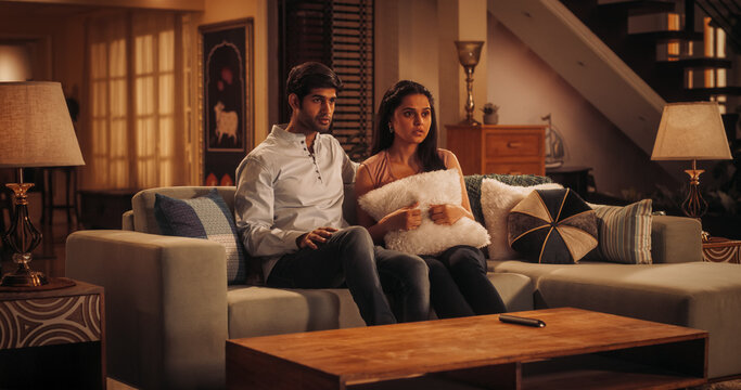 
Young Indian Couple Watching Scary Movie on TV, Finds Comfort In Each Other's Arms As They Sit On Their Couch. Boyfriend and Girlfriend with Expressive Faces Focused on an Exciting Action Film 