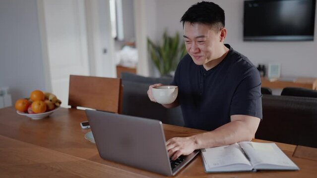 Cheerful asian man working on laptop and drinking coffee at home