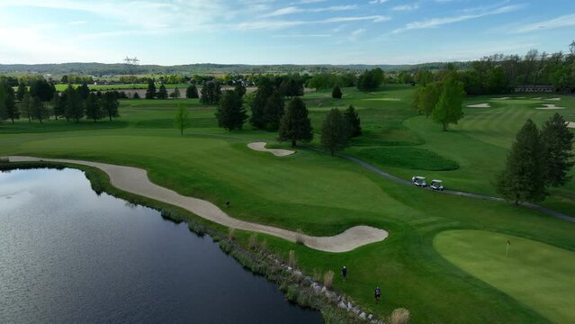 Aerial drone shot of green golf course and pond on a sunny day. Blue sky above rural farmland and manicured golf course. Large sand traps and water hazard featured. Golfers enjoy a day on the green.