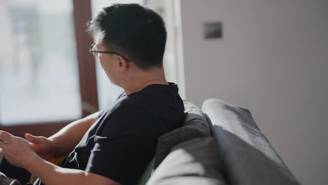 Pretty asian man in eyeglasses looking to side and texting on mobile while sitting at home
