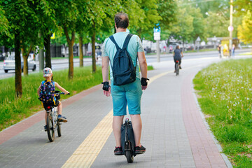 Man on electric unicycle (EUC) riding on bike lane with little son on bicycle, happy family. Father with kid cycling on city street together, sports and leisure outdoor activity.
