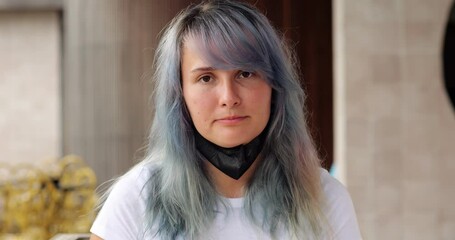Young woman with colored hair wear black mask, portrait shot with blurred background, she look straight in camera. Then girl pull mask down to open nose and mouth and make sigh