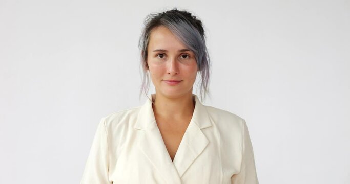 Woman look straight to the camera and smile, we see dental braces on her teeth, then she close lips. Young lady wear light jacket and have colored hair, portrait shot against white background