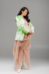 full length of pregnant woman with wavy brunette hair posing in green and white blazer, crop top and beige chiffon cloth on grey background, maternity fashion concept, expectation