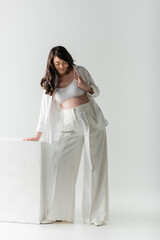 full length of pleased pregnant woman with wavy brunette hair, wearing white pants, crop top and shirt, standing near cube on grey background, maternity fashion concept