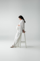 side view of mother-to-be with wavy brunette hair posing near stool in white pants, crop top and...