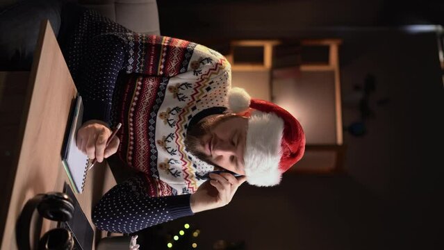 Male freelancer in Santa hat answering phone call and writing in notebook while working on remote project on Christmas day at home