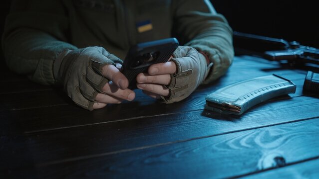A Ukrainian soldier sits at a wooden table, on which lies his military weapon and ammunition. Close up of male hands holding a smartphone. Letter from the war.