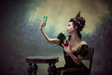 Portrait of young beautiful girl, princess in elegant dress taking selfie with mobile phone and drinking red wine over dark vintage background. Concept of history, renaissance art, comparison of eras